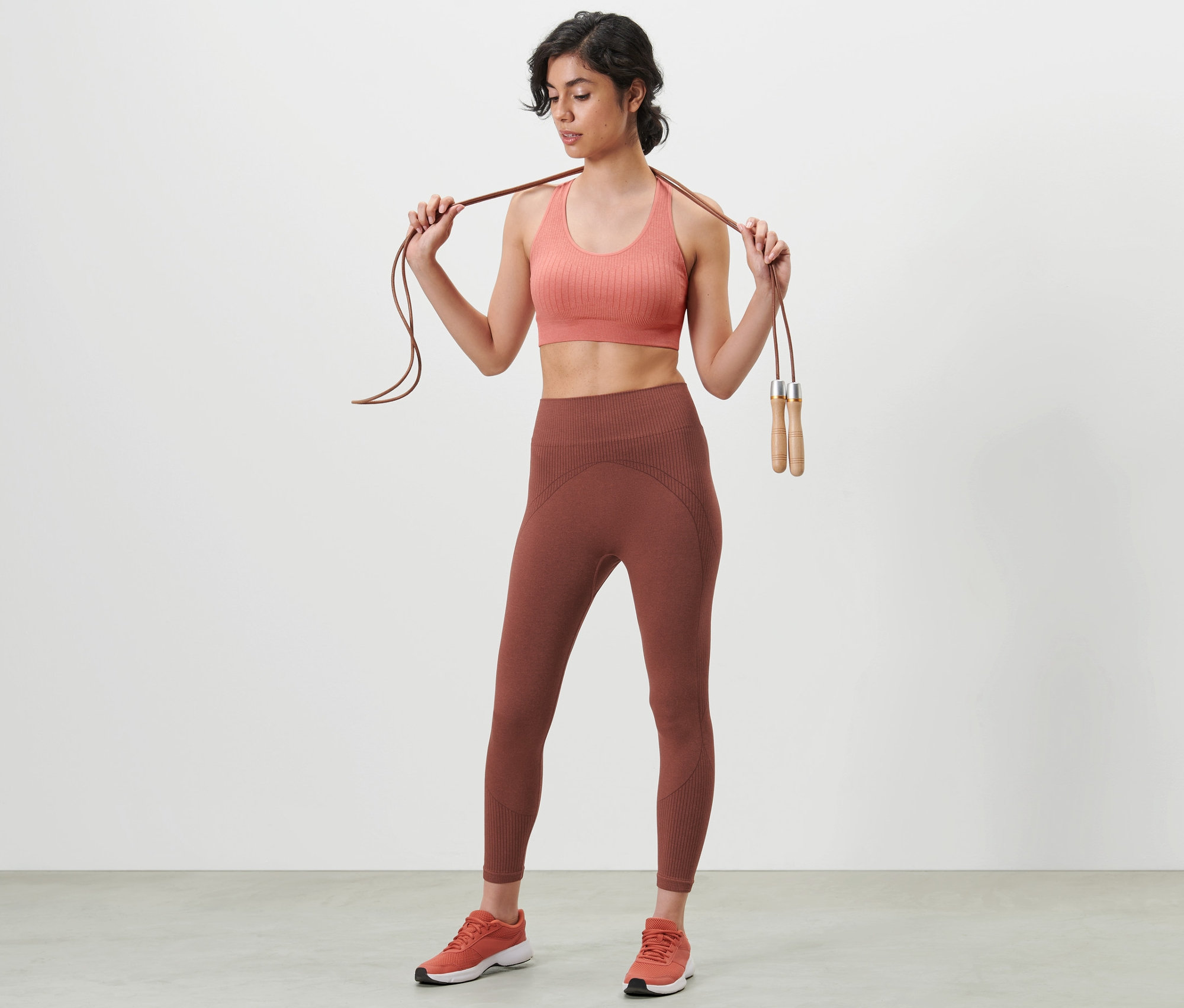 Kobieta trzyma skakankę na ramionach, ma na sobie biustonosz sportowy i bezszwowe legginsy funkcyjne o długości 7/8.