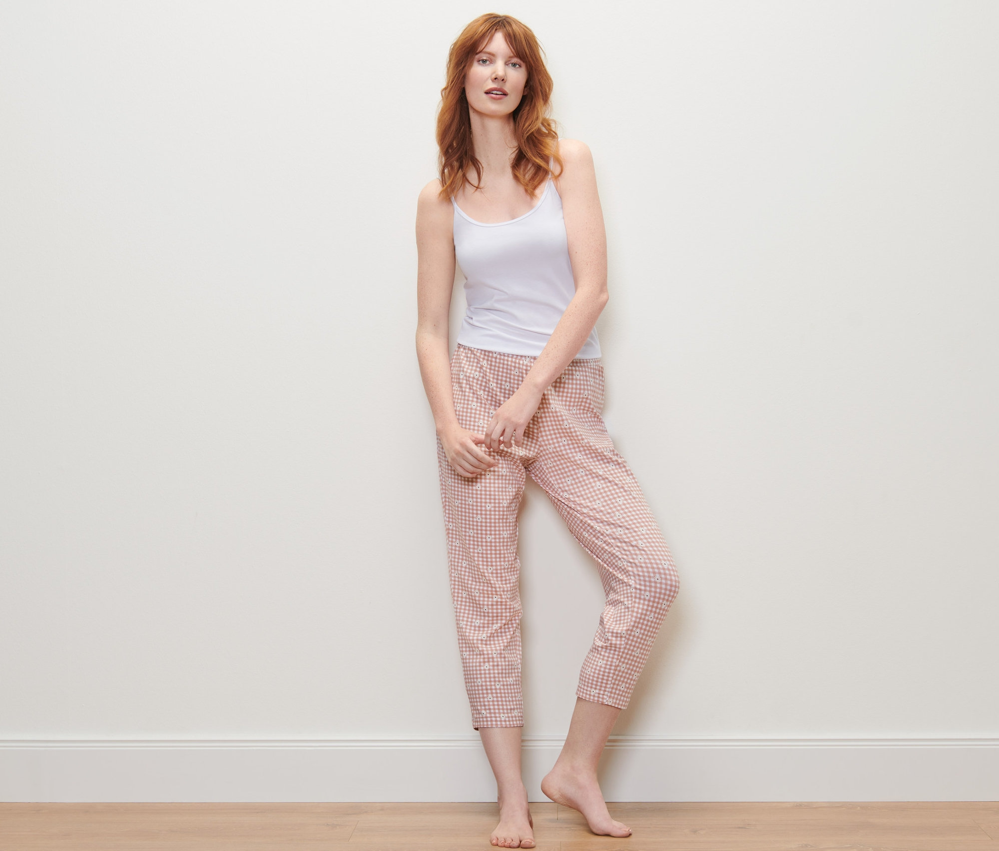 Ruda kobieta stoi w białym topie i spodniach od piżamy o długości 7/8 w kratę.