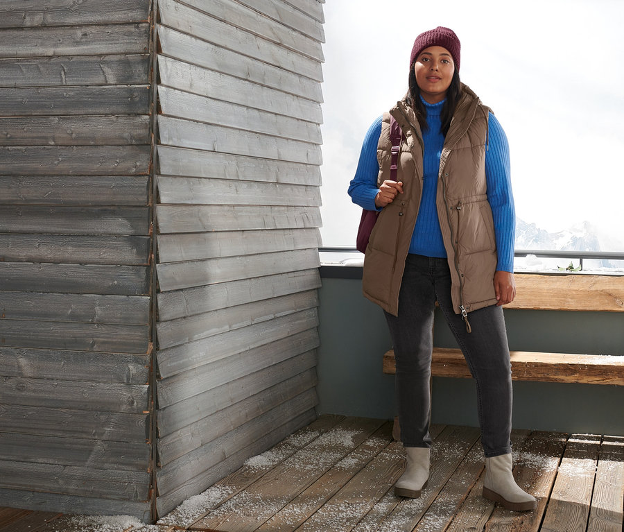 Kobieta stoi na zewnątrz i ma na sobie kamizelkę pikowaną, golf z dzianiny o splocie ściągaczowym, spodnie dżinsowe o kroju slimfit, botki ze skóry zamszowej i bordową czapkę z dzianiny. Ma na sobie pikowany plecak.