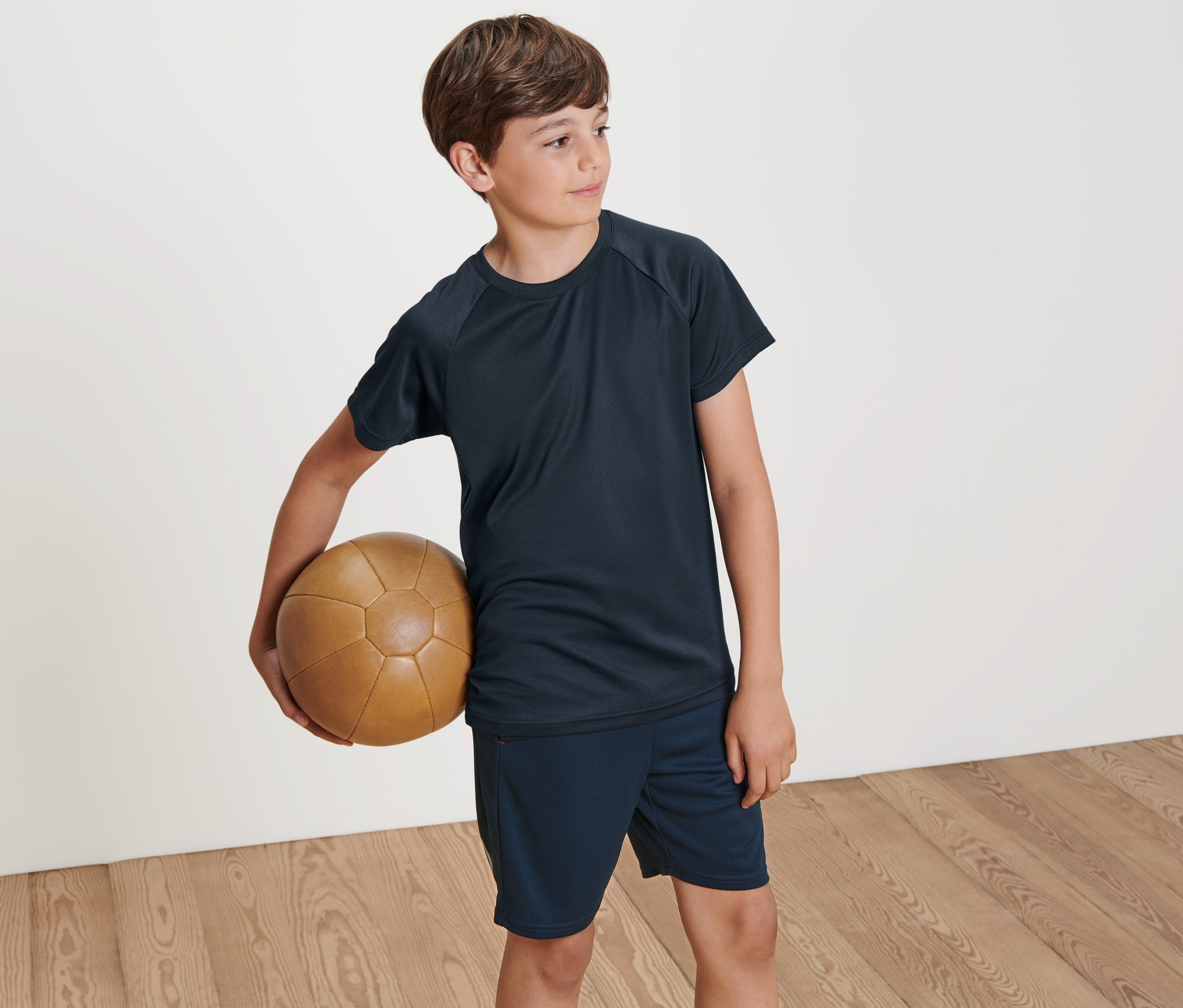 Chłopiec trzyma brązową piłkę skórzaną. Ma na sobie jedną z dwóch koszulek funkcyjnych.