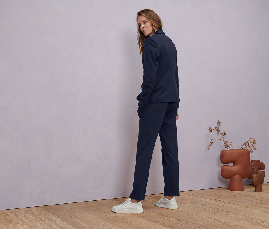 Kobieta spogląda przez ramię i ma na sobie granatowy żakiet z dżerseju, granatowe spodnie dżersejowe z kantem i białą bluzkę z półdługim rękawem.