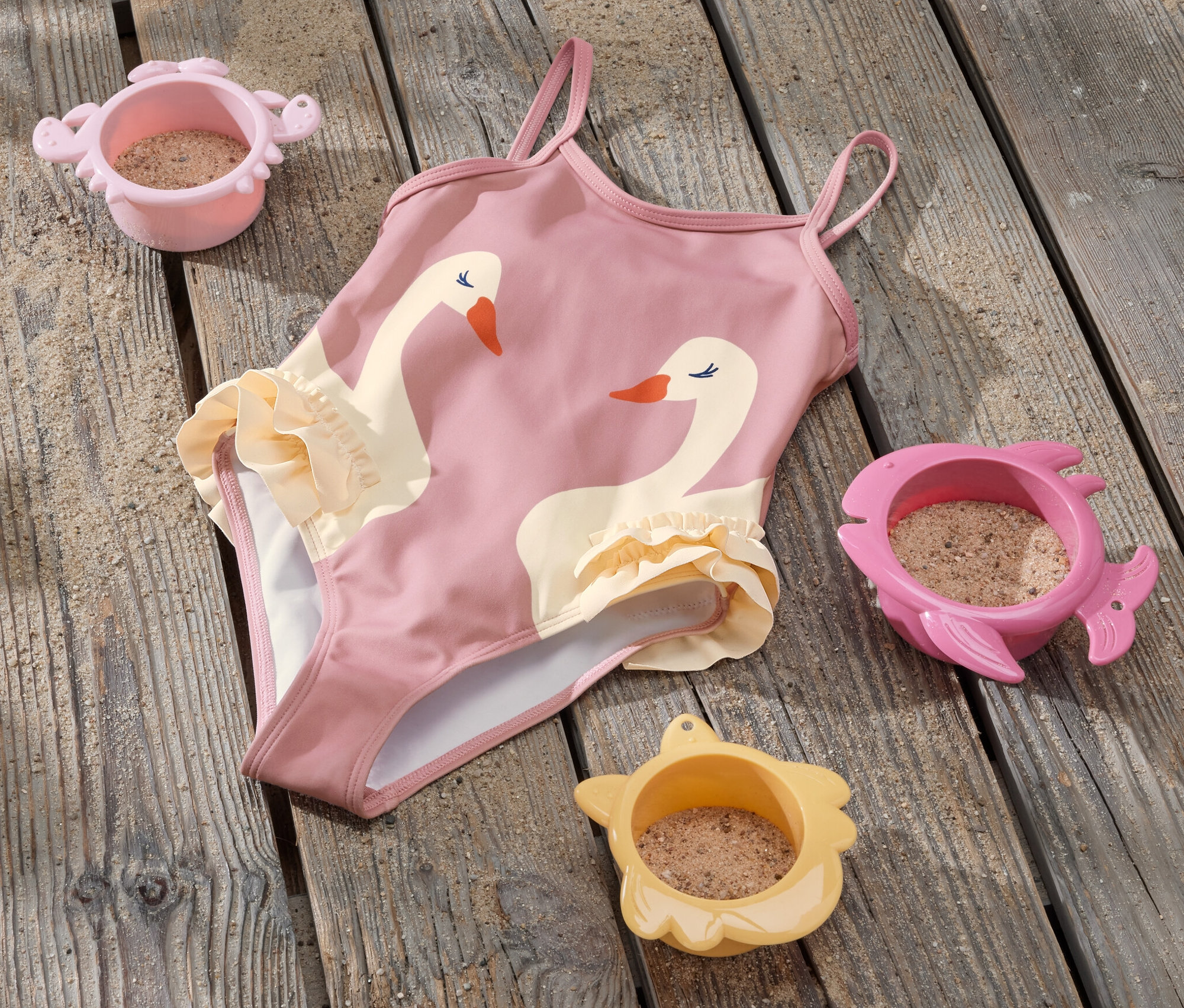 Kostium kąpielowy z falbankami i motywem łabędzia leży na drewnianej podłodze obok trzech zabawek do piasku.