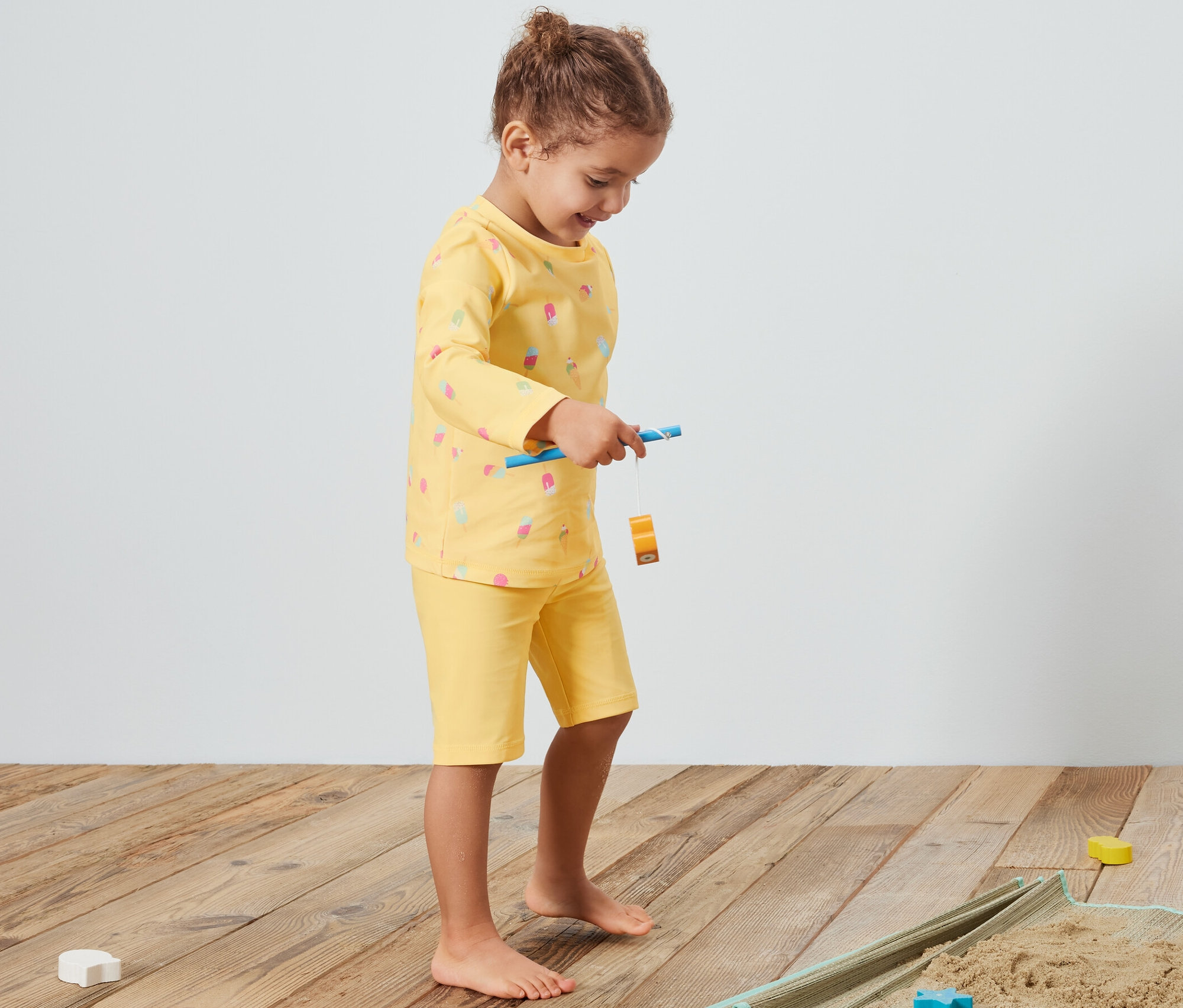 Dziewczynka bawi się na kocu z piaskiem, ubrana w żółty komplet z filtrem UV 80.