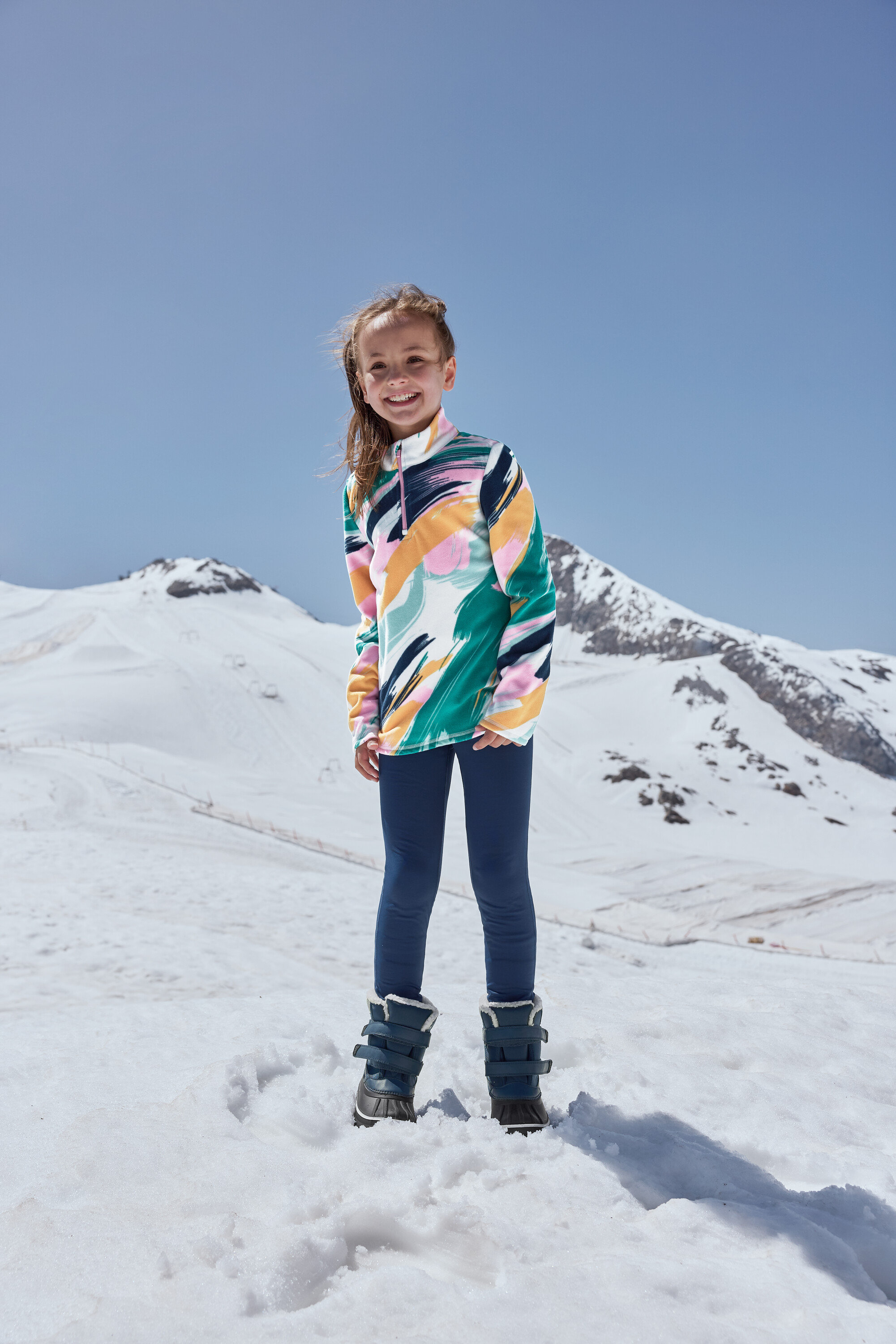 Uśmiechnięta dziewczyna stoi na śniegu w niebieskich dziecięcych butach na każdą pogodę.