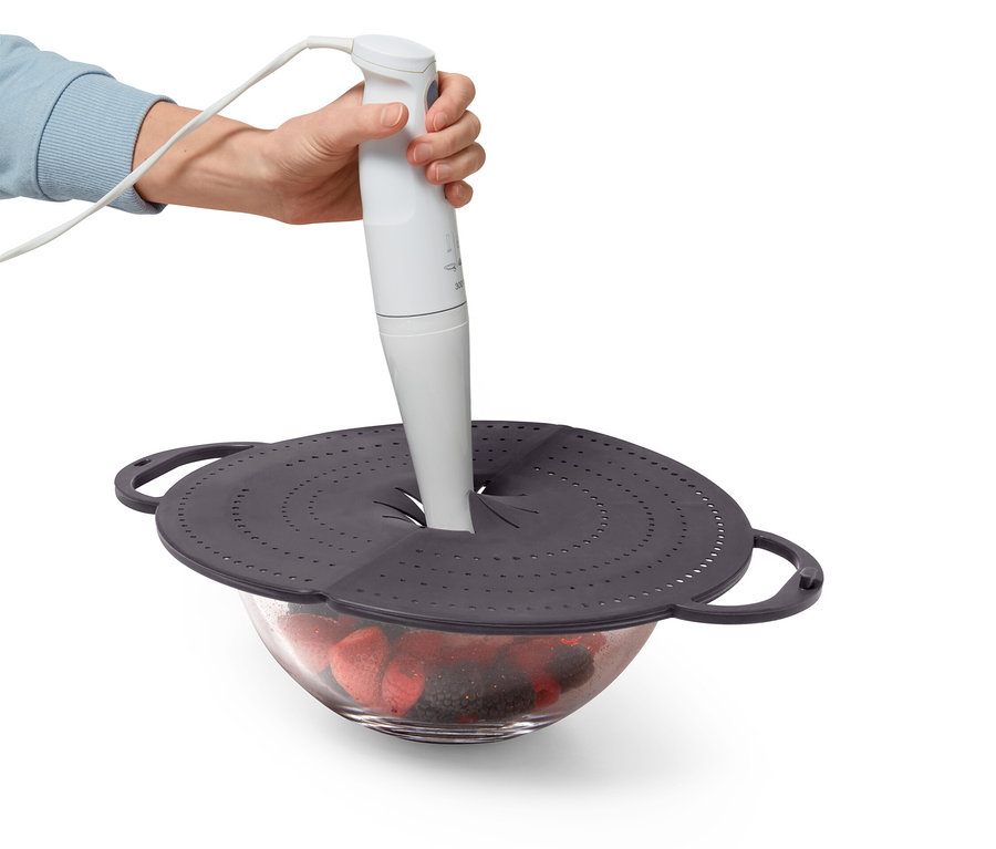 Ręka trzyma biały blender nad miską z owocami, która jest przykryta szarą pokrywką z otworami.