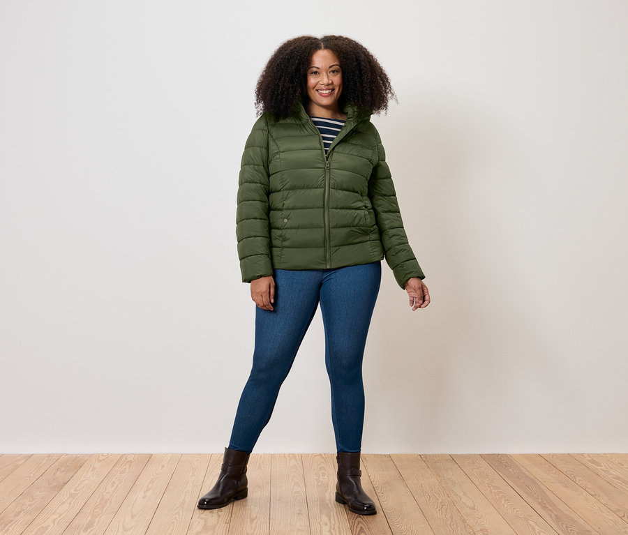 Kobieta stoi, ubrana w oliwkową kurtkę puchową, dżinsy i brązowe buty.