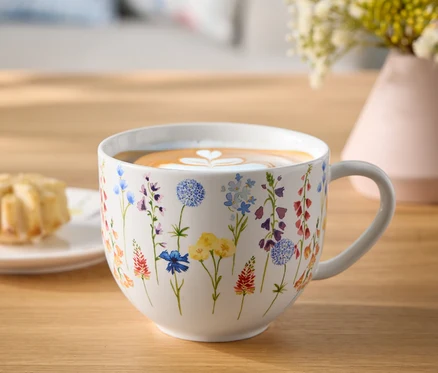 Kubek w kwiaty z kawą i latte art w kształcie serca na drewnianym stole obok talerza z ciastem i wazonu z kwiatami.