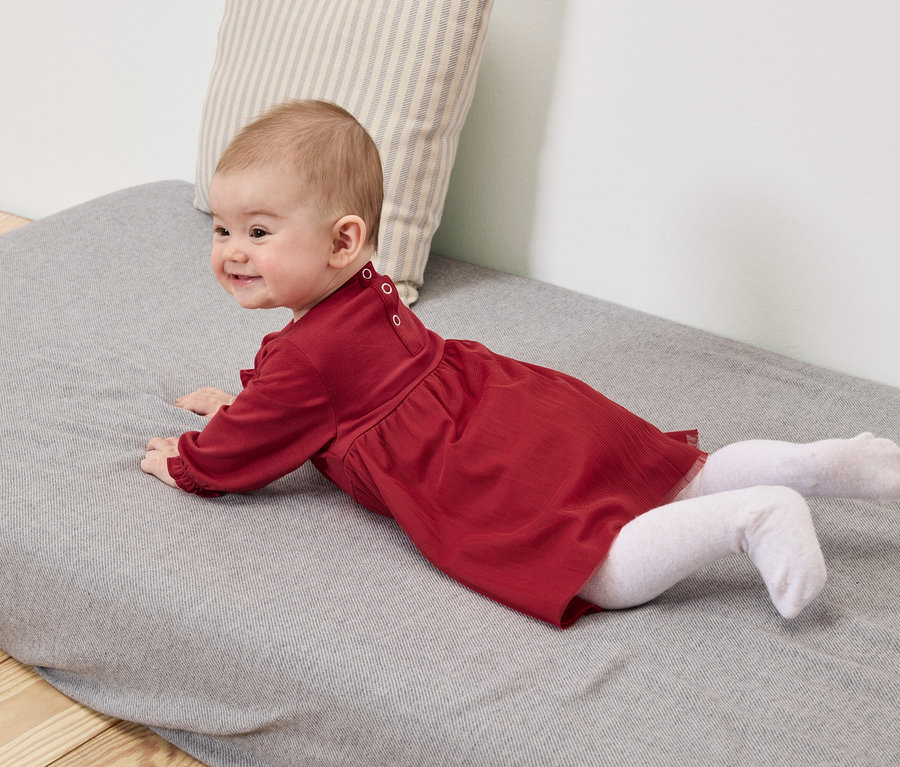 Dziecko w czerwonej sukience i białych rajstopach leży na szarej macie.