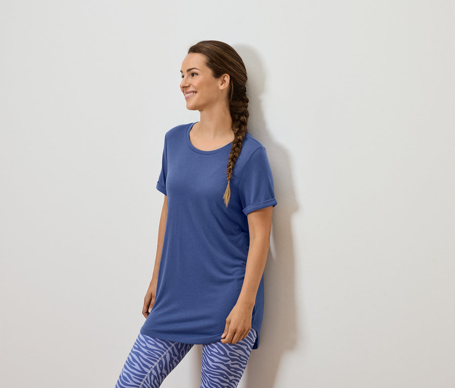 Kobieta z brązowym warkoczem stoi przy białej ścianie, ubrana w niebieską koszulkę i niebieskie legginsy we wzory zwierzęce.