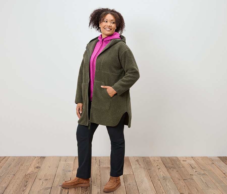 Kobieta w oliwkowym płaszczu z kapturem, różowej bluzie z kapturem, granatowych spodniach i brązowych butach stoi na drewnianej podłodze.