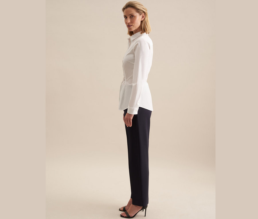 Kobieta stoi bokiem, ubrana w białą koszulę slim fit Seidensticker i granatowe spodnie.