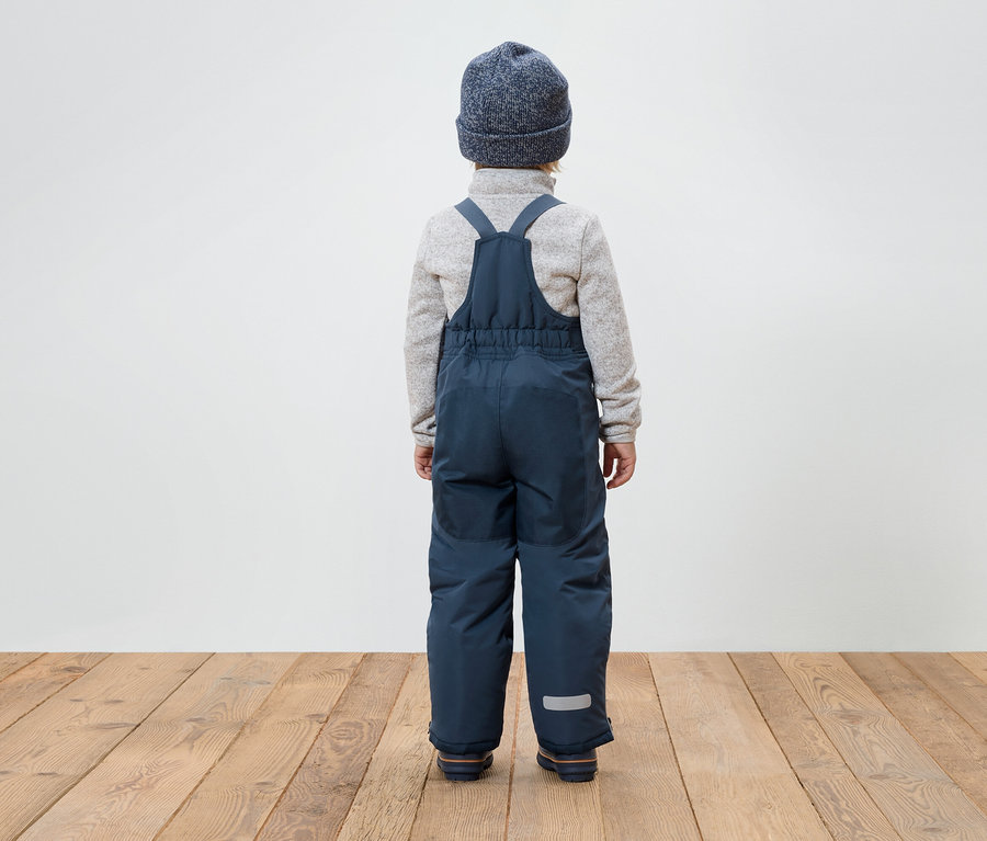 Chłopiec tyłem do kamery ma na sobie niebieskie spodnie na szelkach, szary sweter i niebieską czapkę.