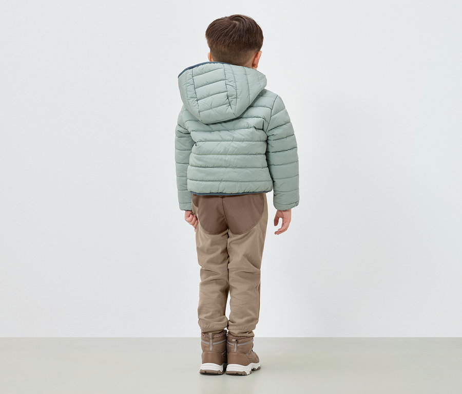Chłopiec stoi tyłem, ubrany w jasnozieloną puchową kurtkę z kapturem, brązowe spodnie i brązowe buty.