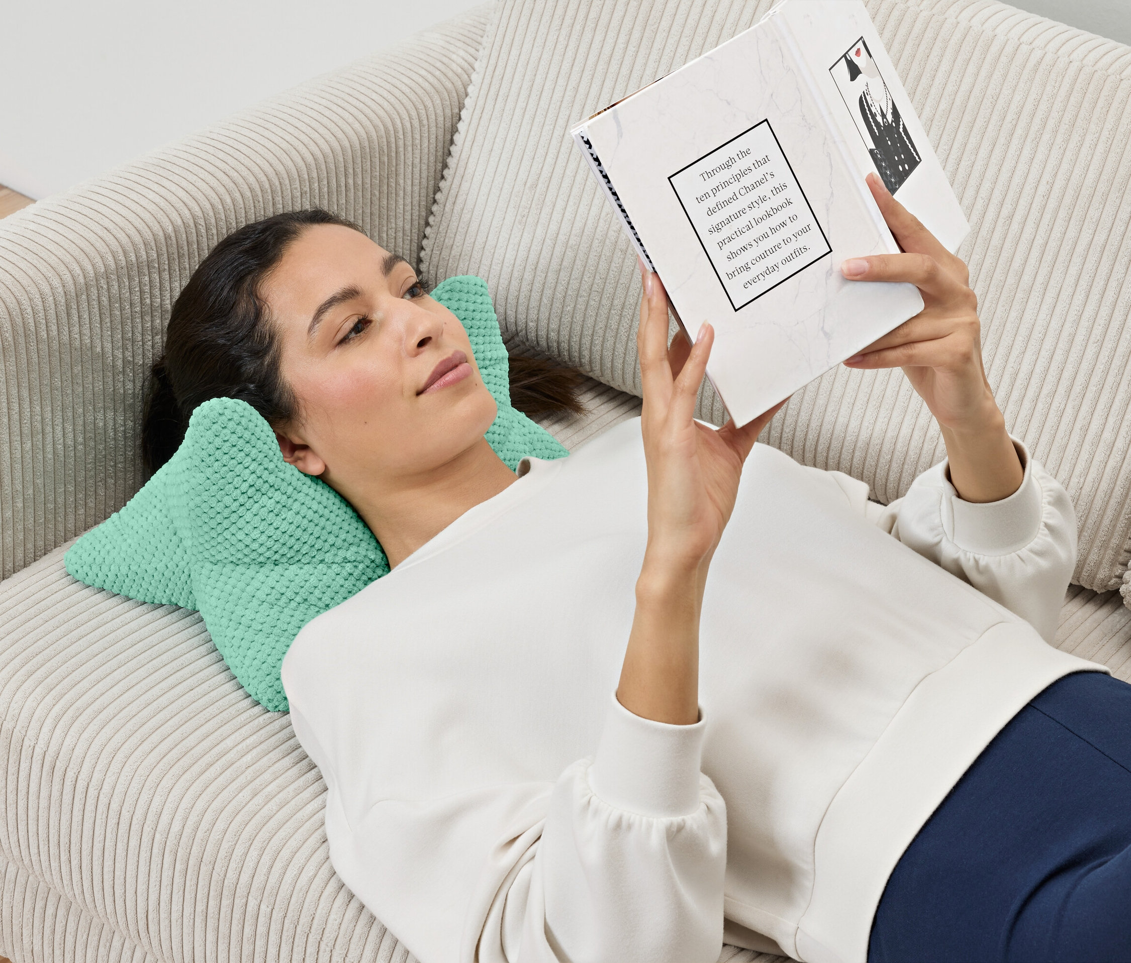 Kobieta leży na sofie i czyta książkę, opierając głowę na poduszce do czytania.