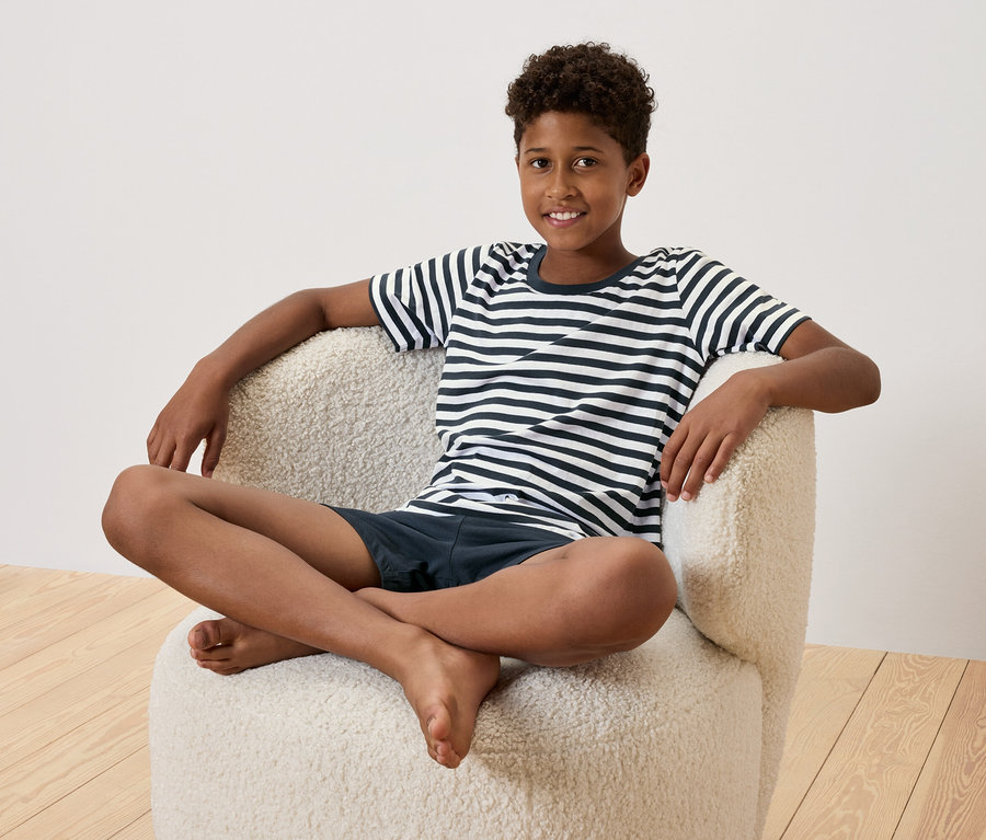 Młody chłopak siedzi ze skrzyżowanymi nogami w kremowym fotelu. Ma na sobie koszulkę w paski i granatowe szorty.