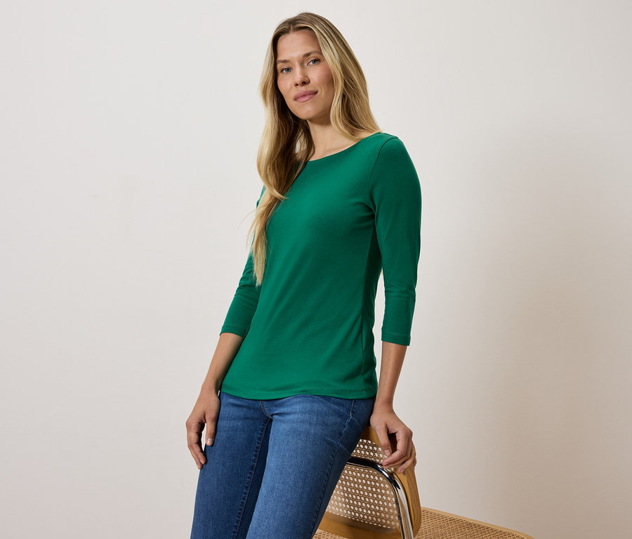 Blondynka w zielonej koszulce z rękawem trzy czwarte i niebieskich dżinsach siedzi na krześle.