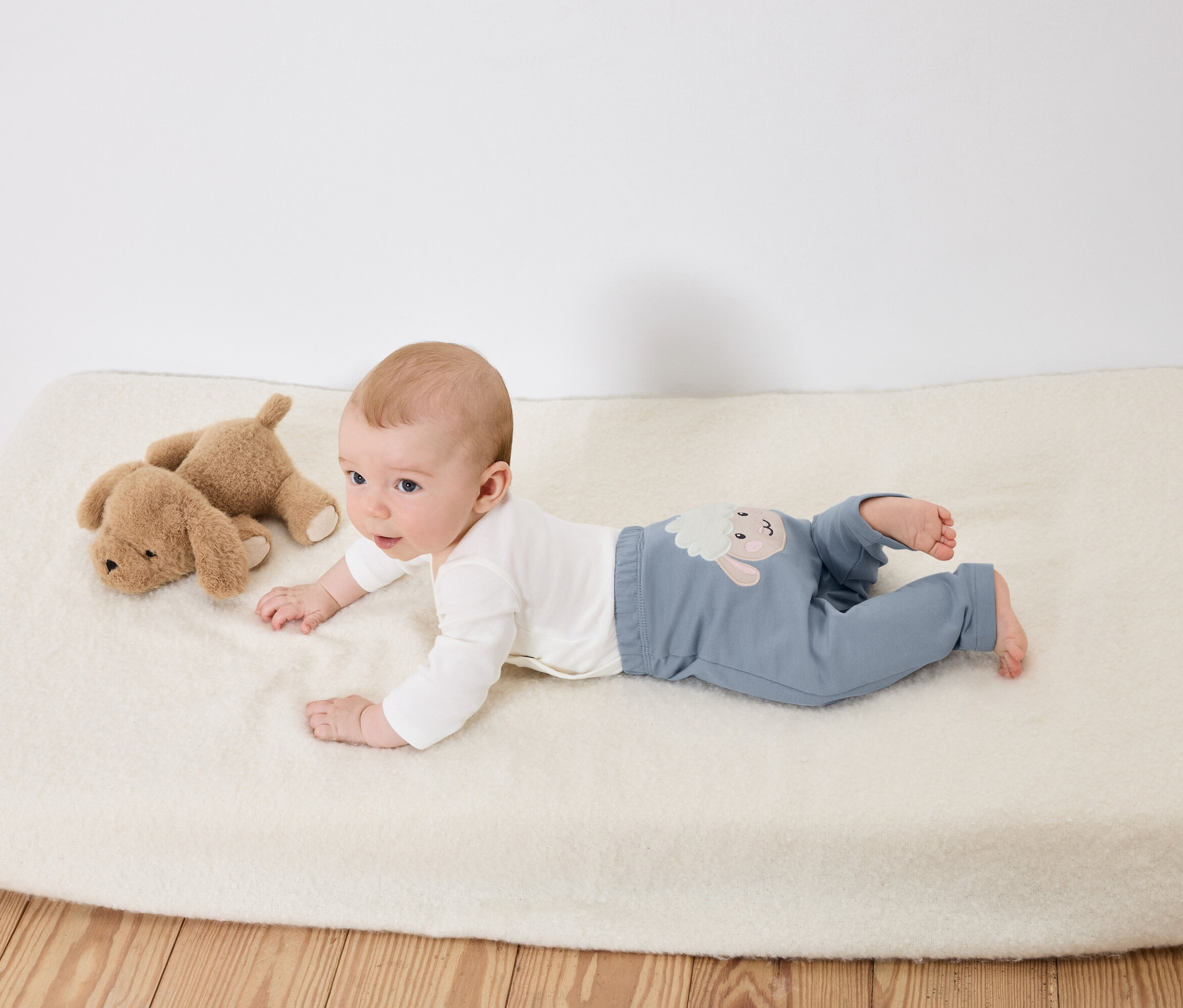 Niemowlę leży na brzuchu na białej macie obok dwóch pluszowych psów.