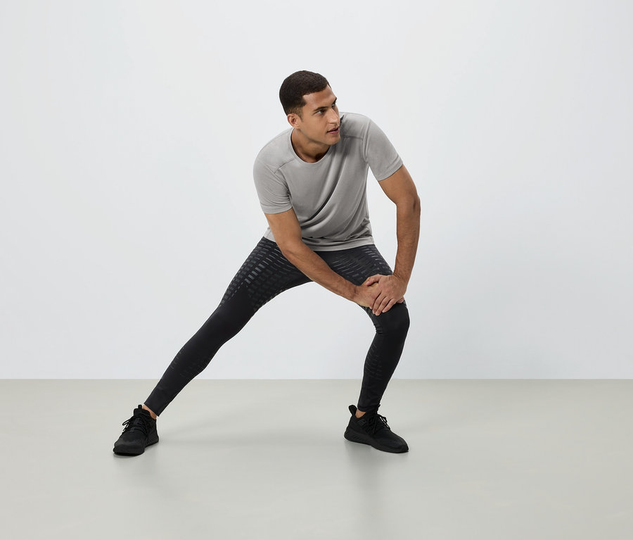 Mężczyzna rozciąga się w szarej koszulce, czarnych legginsach i czarnych butach sportowych na białym tle.