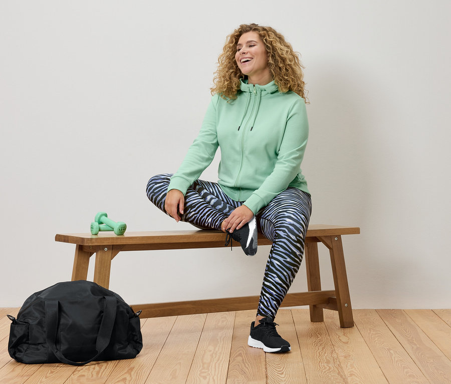 Kobieta z kręconymi włosami siedzi na drewnianej ławce, ubrana w miętową bluzę z kapturem i legginsy w zebrę. Widoczna jest czarna torba sportowa i zielone hantle.
