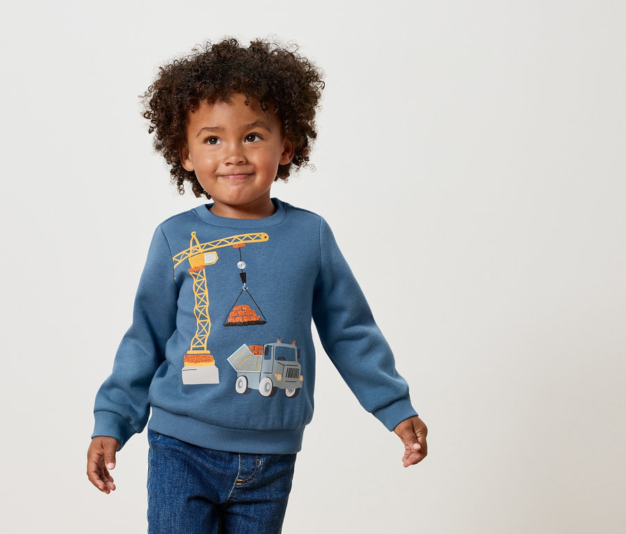 Chłopiec z kręconymi włosami ma na sobie niebieską bluzę z aplikacją przedstawiającą żółty dźwig i ciężarówkę.