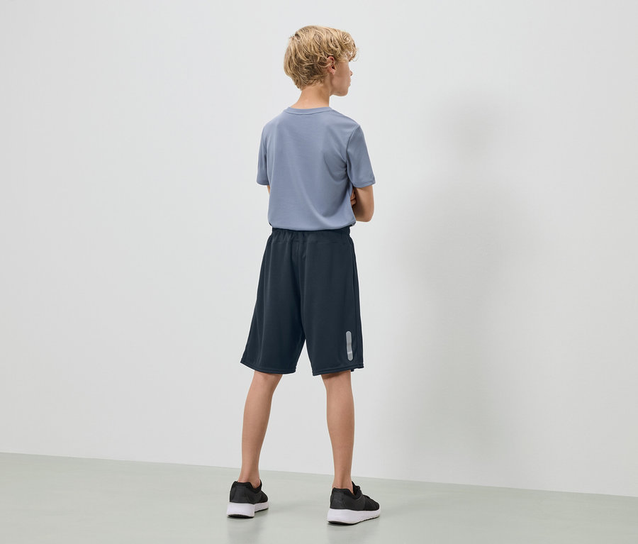 Nastolatek z blond, kręconymi włosami stoi tyłem do kamery, ma na sobie niebieską koszulkę, granatowe szorty i czarne tenisówki.