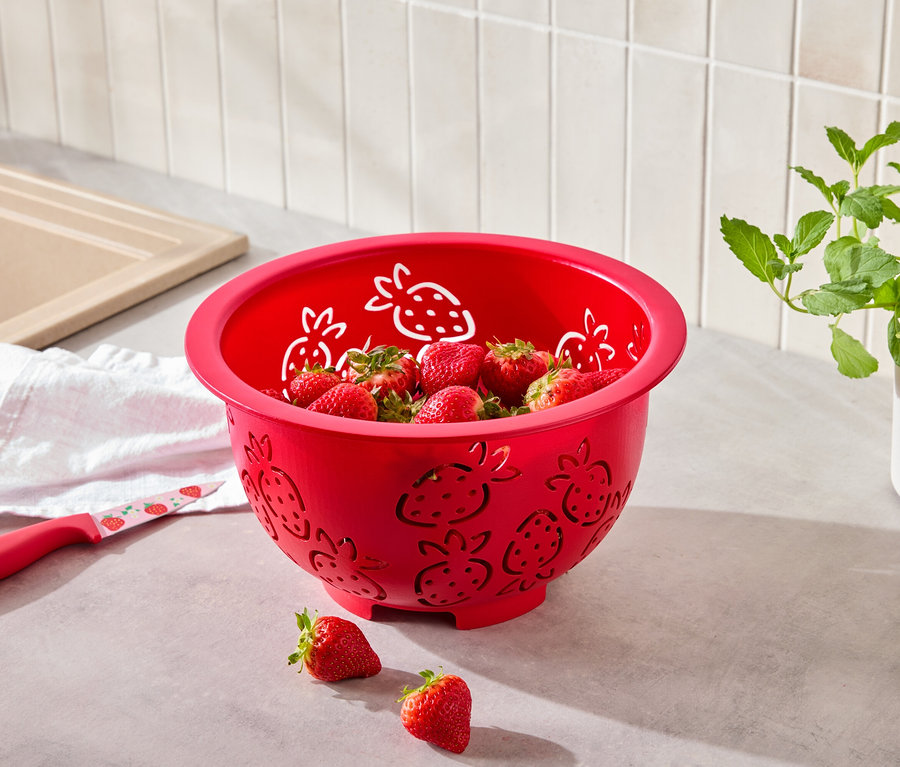 Czerwony durszlak z motywem truskawek wypełniony truskawkami, obok leży nóż z motywem truskawek.