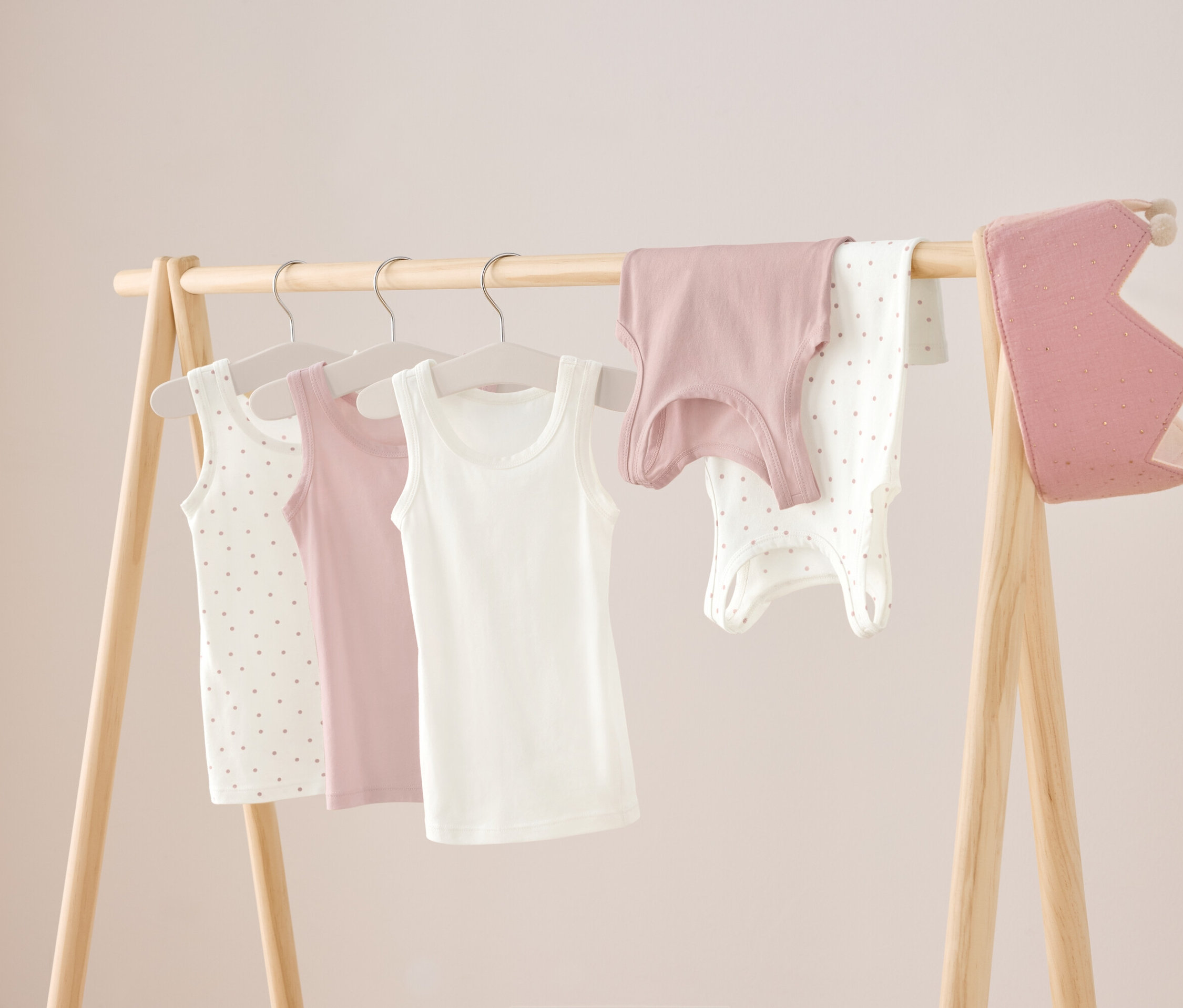 Na drewnianym stojaku wiszą trzy podkoszulki: biały, różowy i biały w kropki, a także różowa koszulka i białe body w kropki. Po prawej stronie wisi różowa korona.