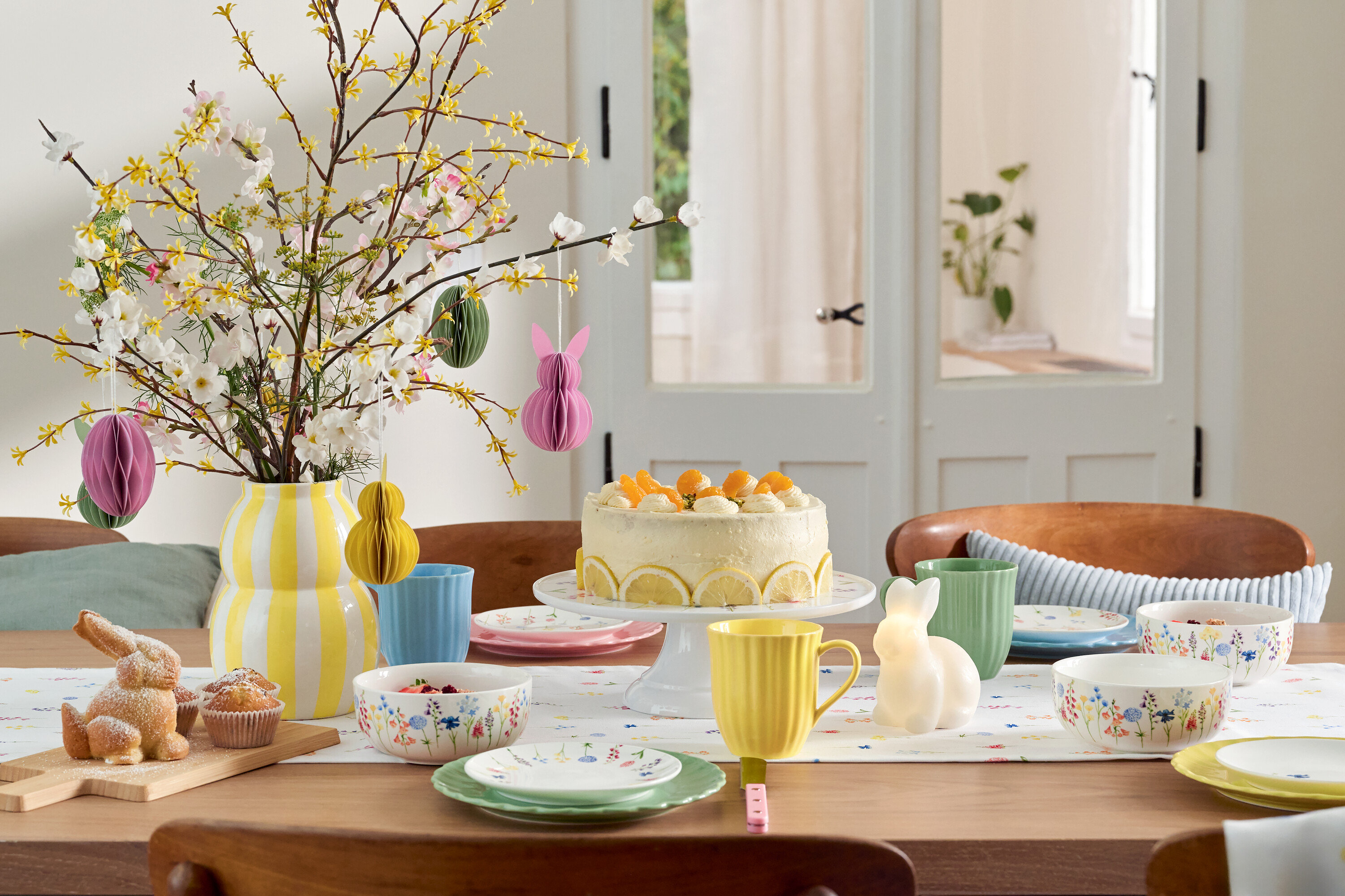 Stół wielkanocny z ciastem, naczyniami i wazonem z kwiatami.