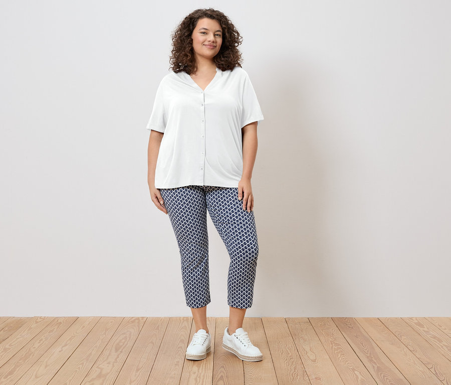 Kobieta o kręconych włosach ma na sobie białą bluzkę koszulową i niebieskie wzorzyste spodnie oraz białe buty.