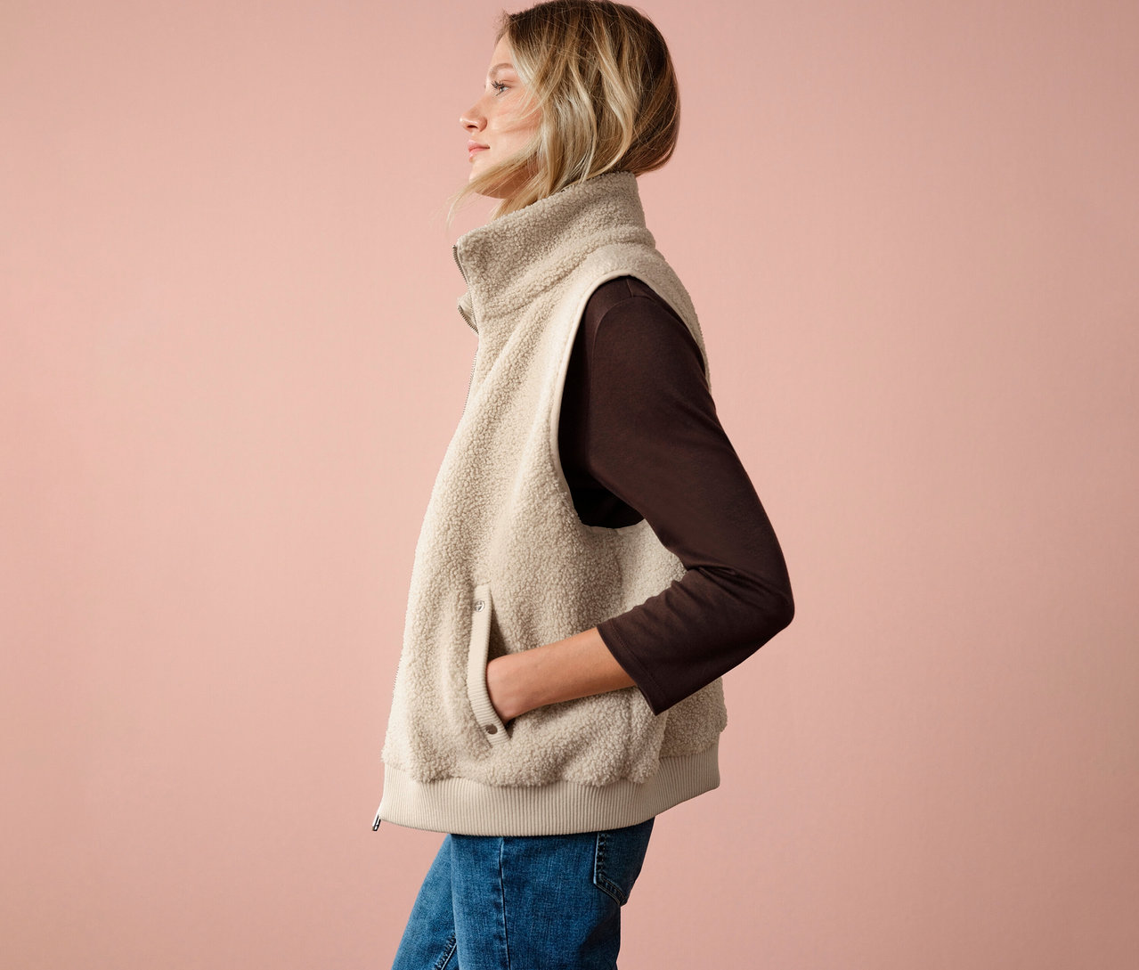 Kobieta w pluszowej kamizelce od Street One, brązowej bluzce i dżinsach, widok z boku na różowym tle.