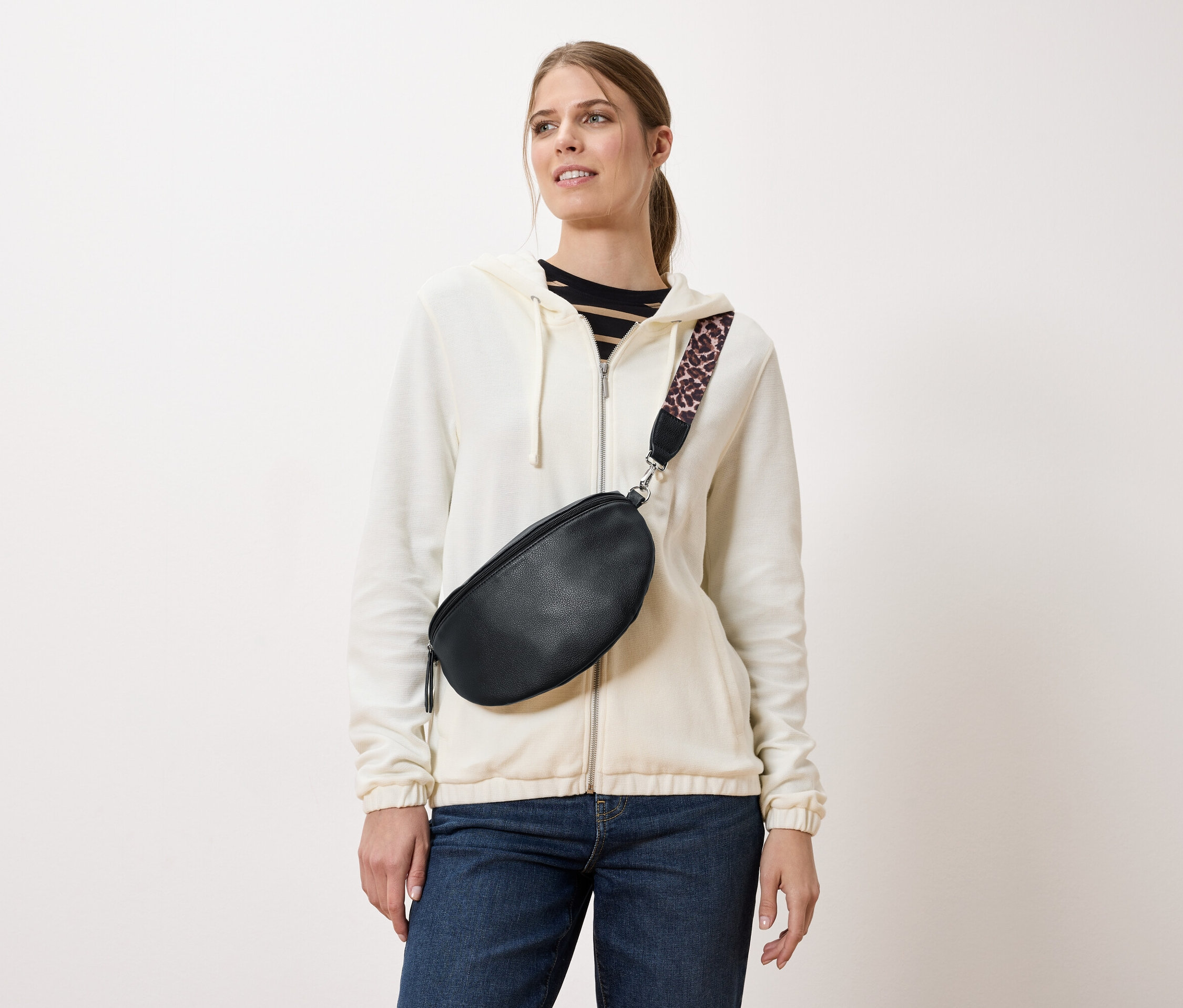 Kobieta nosi czarną saszetkę biodrową z paskiem w lamparcie cętki, założoną na kremową kurtkę i dżinsy.