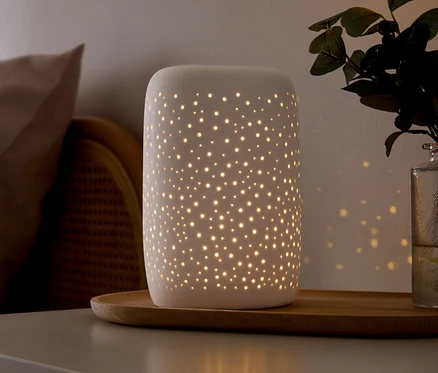 Biała ceramiczna lampa z małymi perforacjami emituje ciepłe światło, rzucając gwiezdne wzory na ścianę. Stoi na drewnianej tacy na szarej powierzchni, obok beżowej poduszki, wiklinowych mebli i zielonej rośliny w szklanym wazonie.