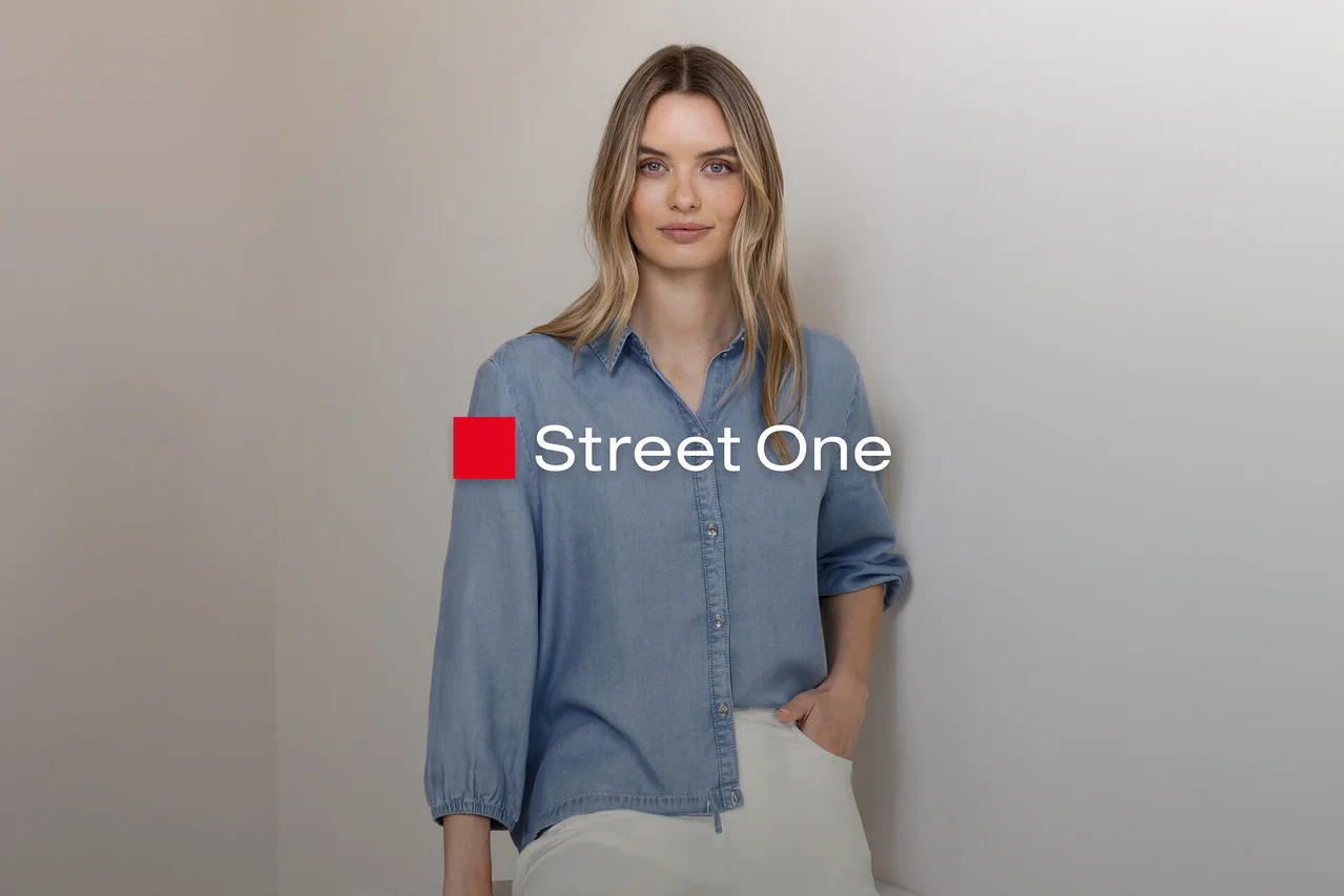 Blondynka w jasnoniebieskiej koszuli dżinsowej i jasnych spodniach, stoi z ręką w kieszeni przy szarej ścianie. Logo Street One.