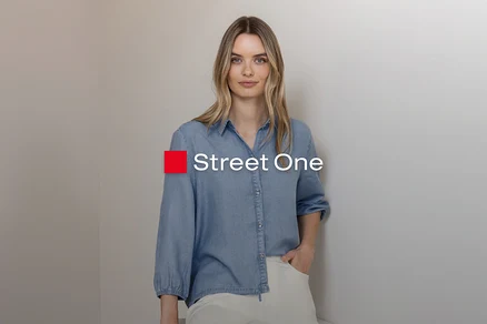 Blondynka w jasnoniebieskiej koszuli dżinsowej i jasnych spodniach, stoi z ręką w kieszeni przy szarej ścianie. Logo Street One.