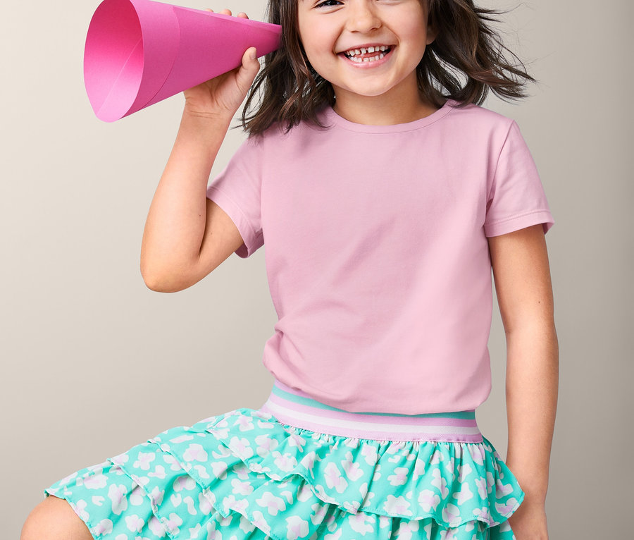 Uśmiechnięta, ciemnowłosa dziewczyna trzyma różowy megafon przy uchu. Ma na sobie koszulki bawełniane, 3 sztuki i turkusową spódnicę.