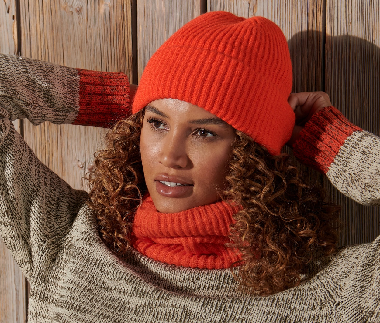 Kobieta z kręconymi włosami pozuje z rękami za głową, ma na sobie pomarańczową czapkę z dzianiny.