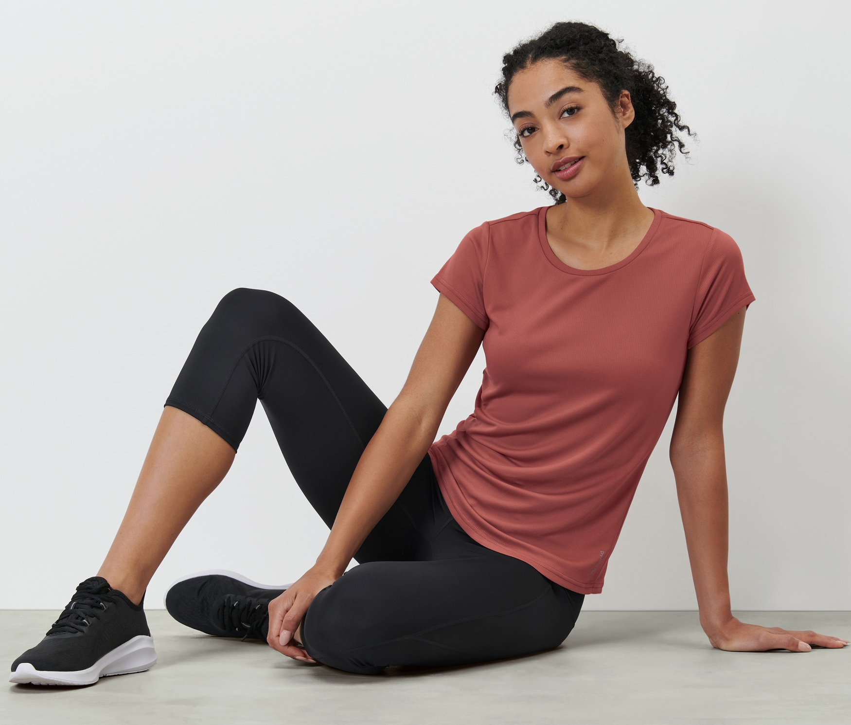 Kobieta siedzi na podłodze w terakotowej koszulce funkcyjnej.