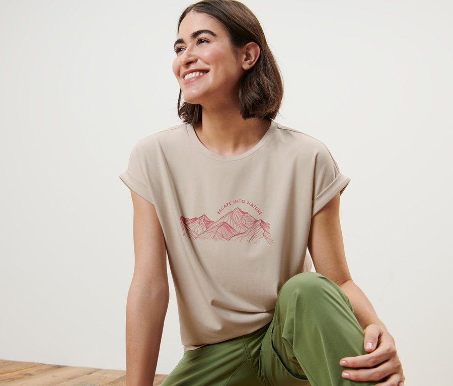 Kobieta siedzi na drewnianej podłodze i ma na sobie beżowy t-shirt z nadrukiem góry.