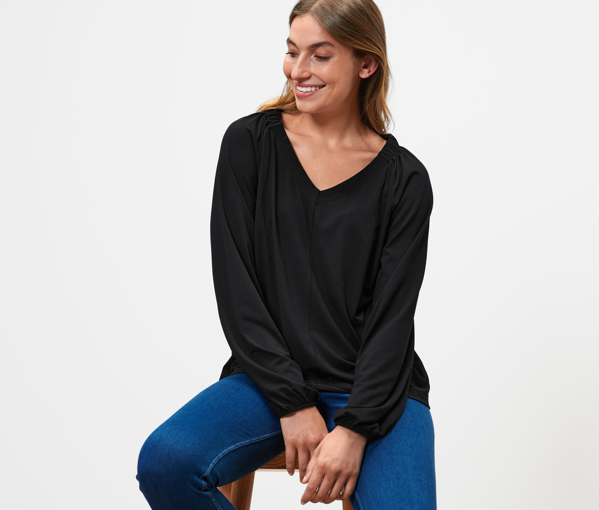 Kobieta siedzi na stołku, ubrana w czarną bluzkę z marszczeniem i niebieskie dżinsy.