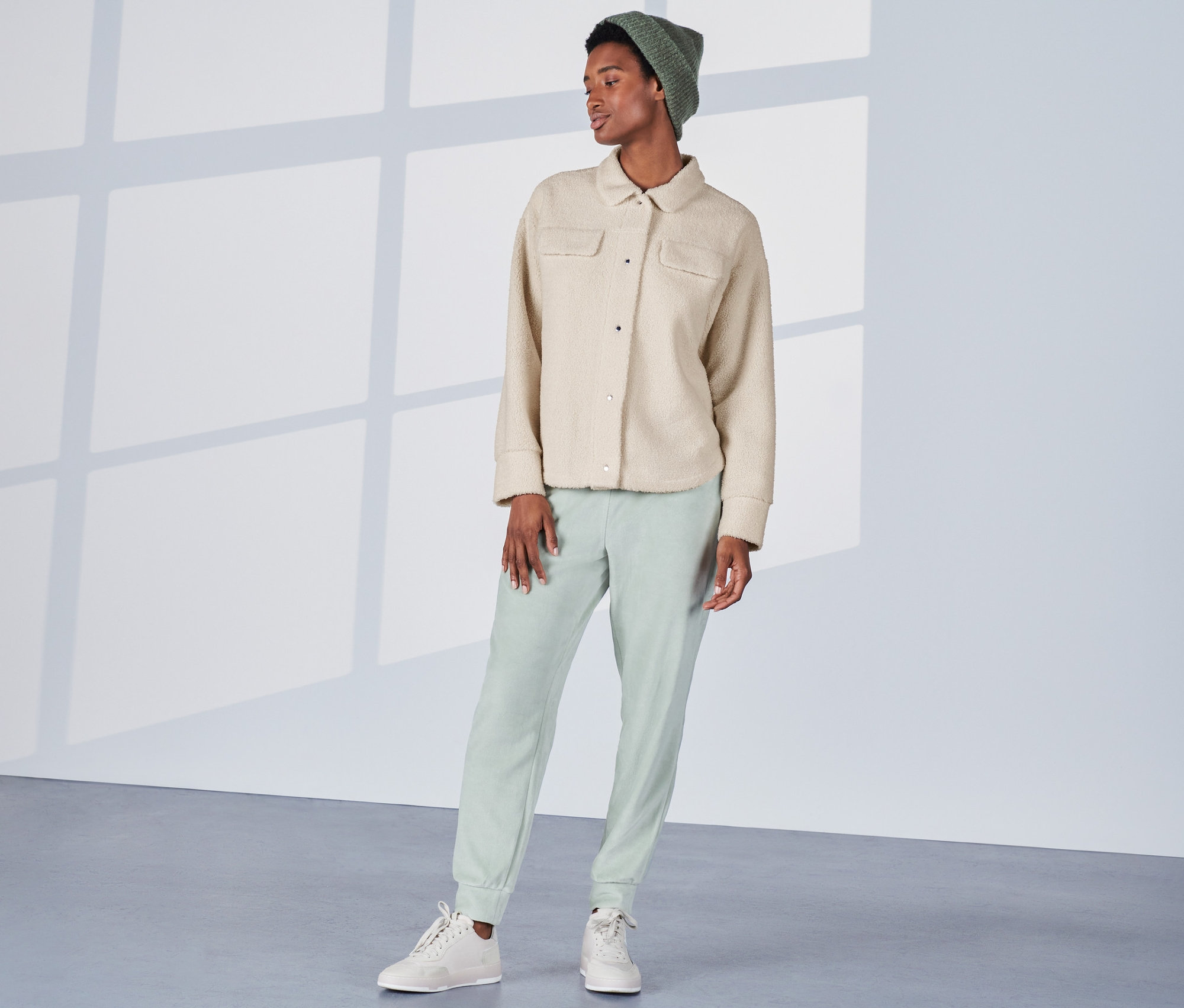 Kobieta stoi i ma na sobie kremową pluszową kurtkę koszulową, zielone spodnie i czapkę.