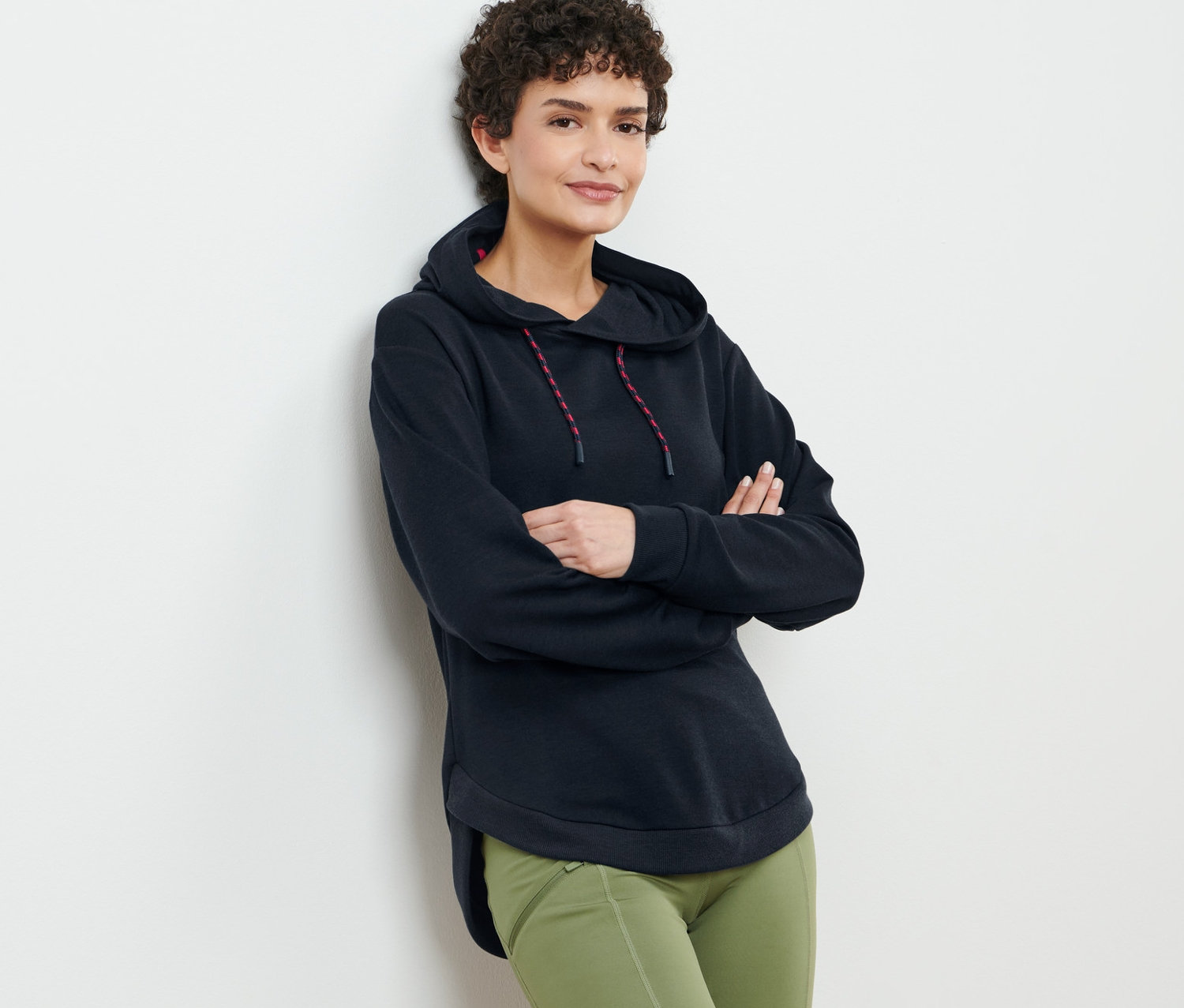 Kobieta o brązowych kręconych włosach pozuje w czarnej bluzie dresowej z kapturem i zielonych spodniach.