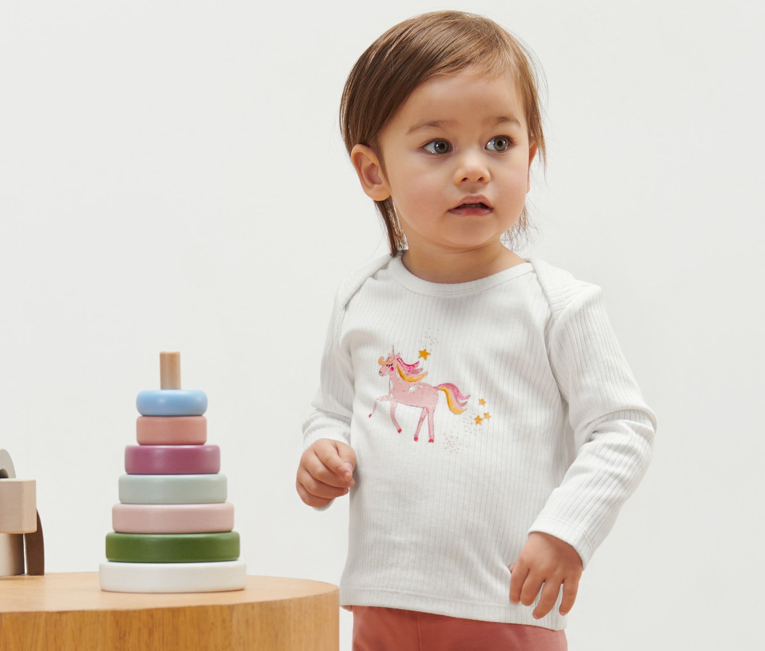 Małe dziecko ma na sobie koszulkę w prążki z jednorożcem. Obok niego stoi drewniana zabawka.