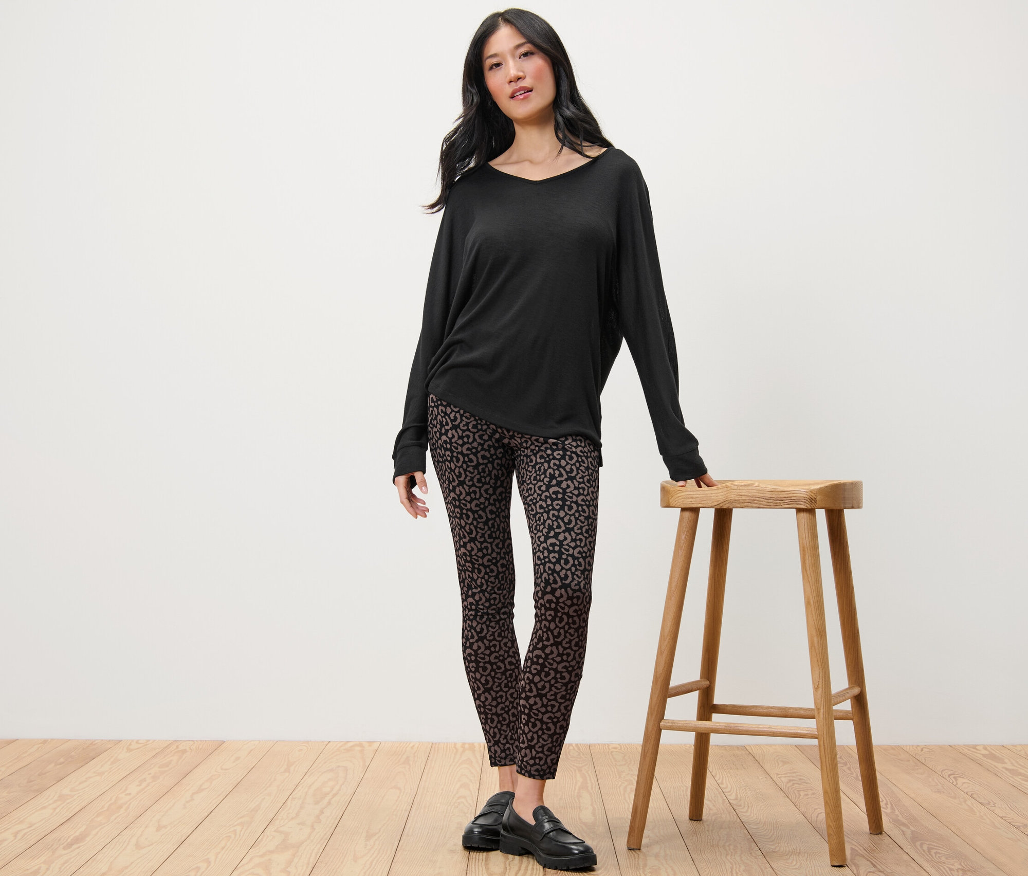 Kobieta stoi obok drewnianego stołka, ma na sobie czarną bluzkę z długim rękawem i grube legginsy.
