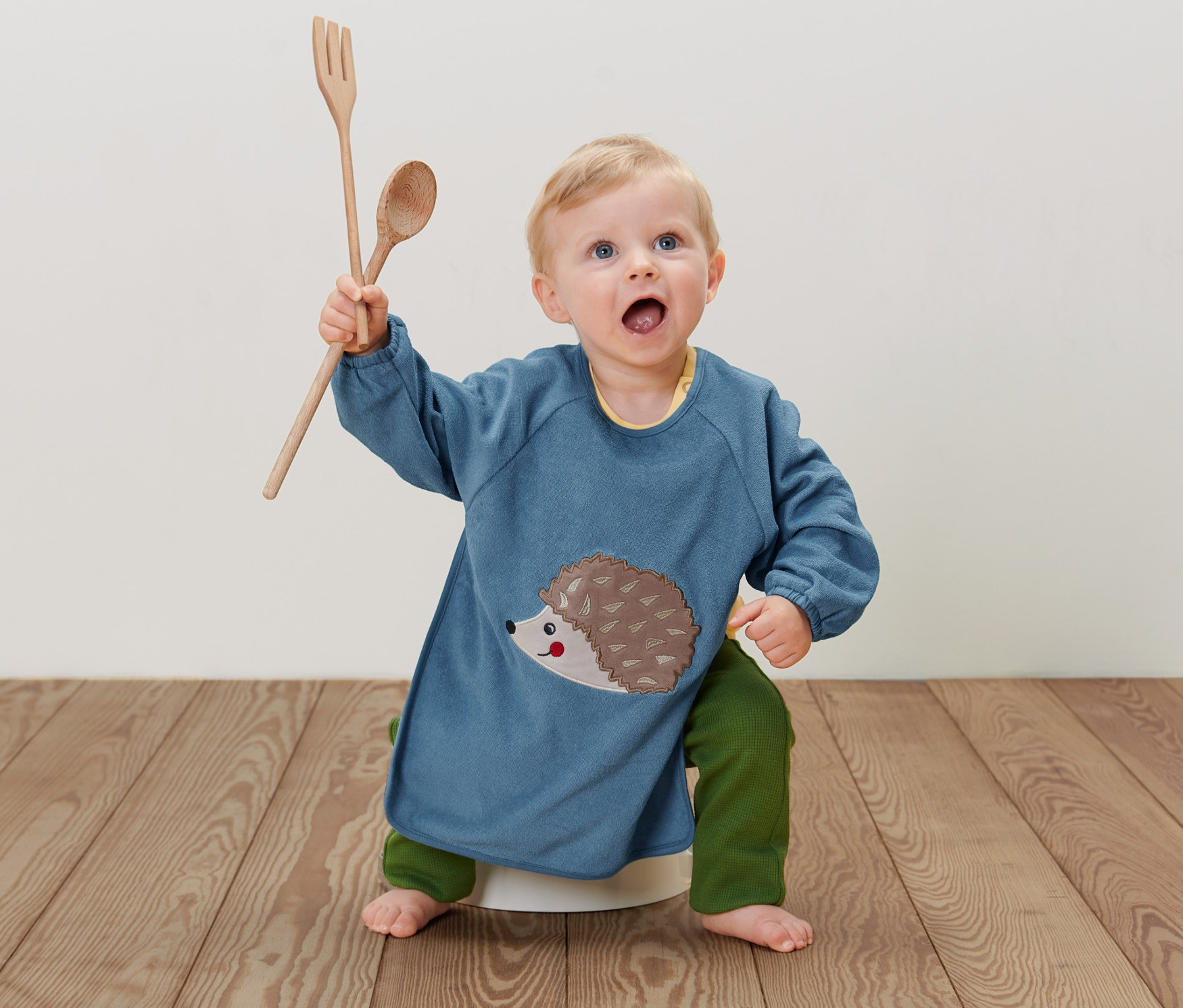 Małe dziecko siedzi na podłodze w niebieskim śliniaku z rękawami, 2 sztuki z jeżem, trzymając drewnianą łyżkę i widelec.