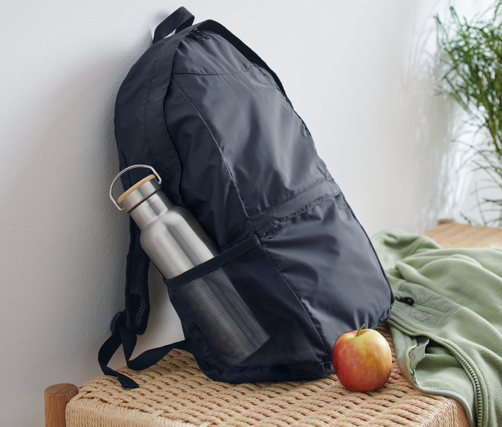 Składany plecak oparty o białą ścianę z butelką wody w bocznej kieszeni i jabłkiem obok na wiklinowej ławce.