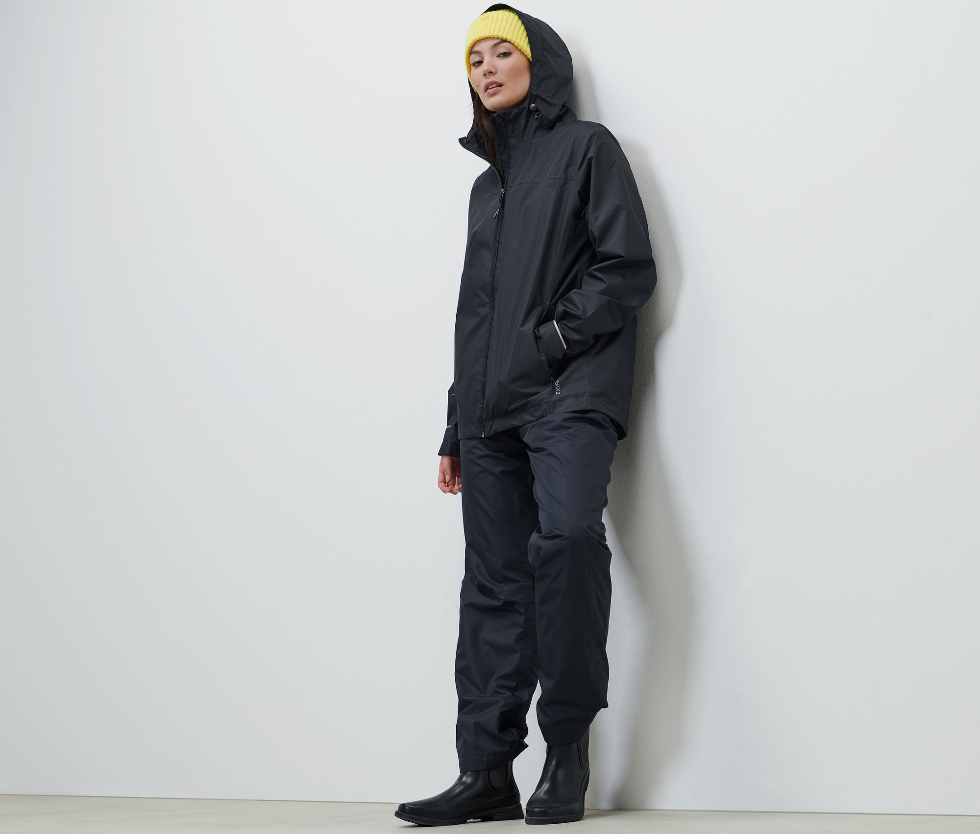 Kobieta ma na sobie granatową kurtkę przeciwdeszczową uniseks, spodnie i żółtą czapkę.