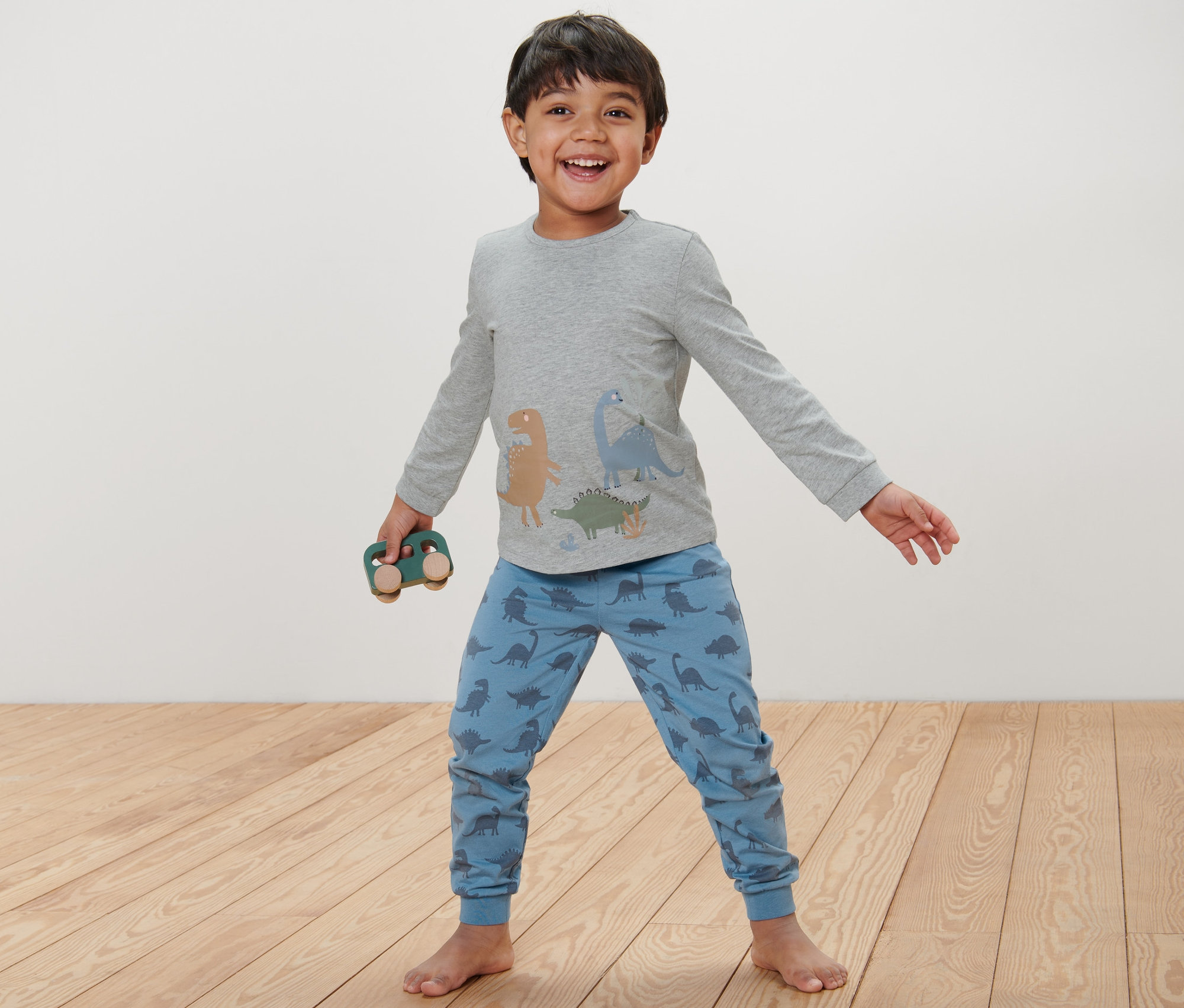 Chłopiec w niebieskiej piżamie dziecięcej w dinozaury trzyma drewniany samochodzik.