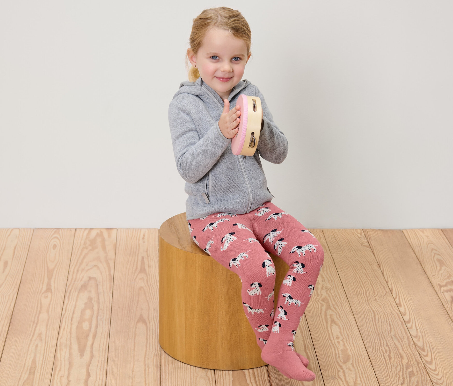 Dziewczynka siedzi i gra na tamburynie, ubrana w różowe i szare dziecięce rajstopy termoaktywne.