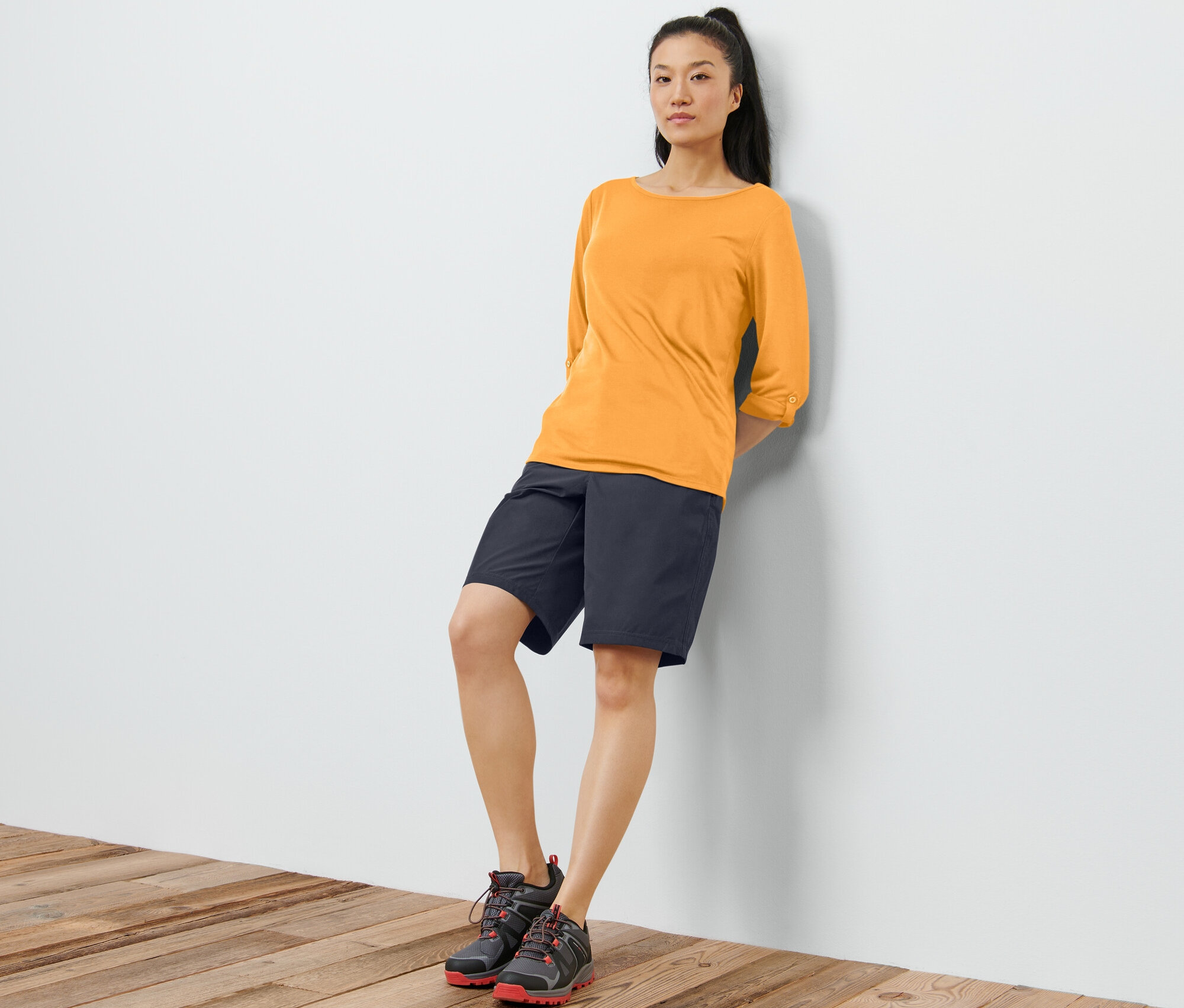 Kobieta opiera się o ścianę, ma na sobie pomarańczową koszulkę funkcyjną z rękawami 3/4, czarne szorty i buty.