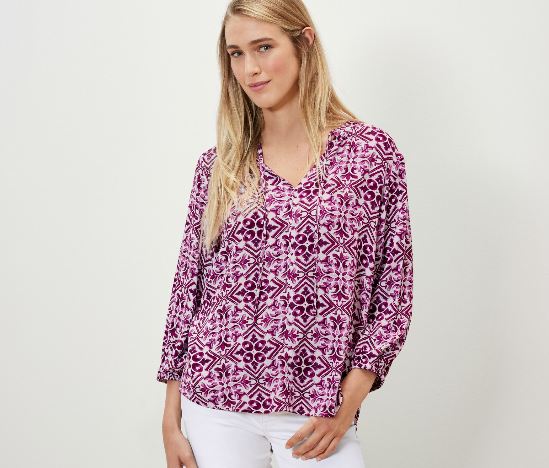 Kobieta ma na sobie bluzkę-tunikę z nadrukiem, jagodową i białe dżinsy.